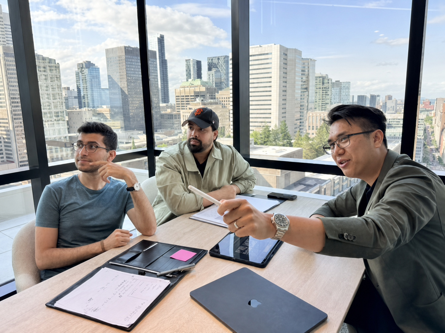 Suti reviewing figures on a tablet with two colleagues at a meeting table, city skyline behind them.